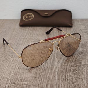 K&ouml;nnte beinhalten: Goldfarbene Sonnenbrille mit braunen Gl&auml;sern und einem Steg in Schildpatt-Optik. Ein braunes Lederetui ist ebenfalls vorhanden. Das Ray-Ban-Logo ist auf dem Glas sichtbar.