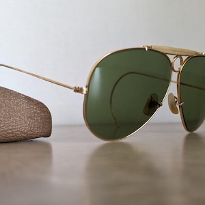 K&ouml;nnte beinhalten: Goldfarbene Aviator-Sonnenbrille mit gr&uuml;nen Gl&auml;sern. Die Sonnenbrille hat einen Doppelsteg und einen Stirnb&uuml;gel. Auf der linken Seite ist ein braunes Lederetui zu sehen. Die Sonnenbrille liegt auf einer hellbraunen Oberfl&auml;che.