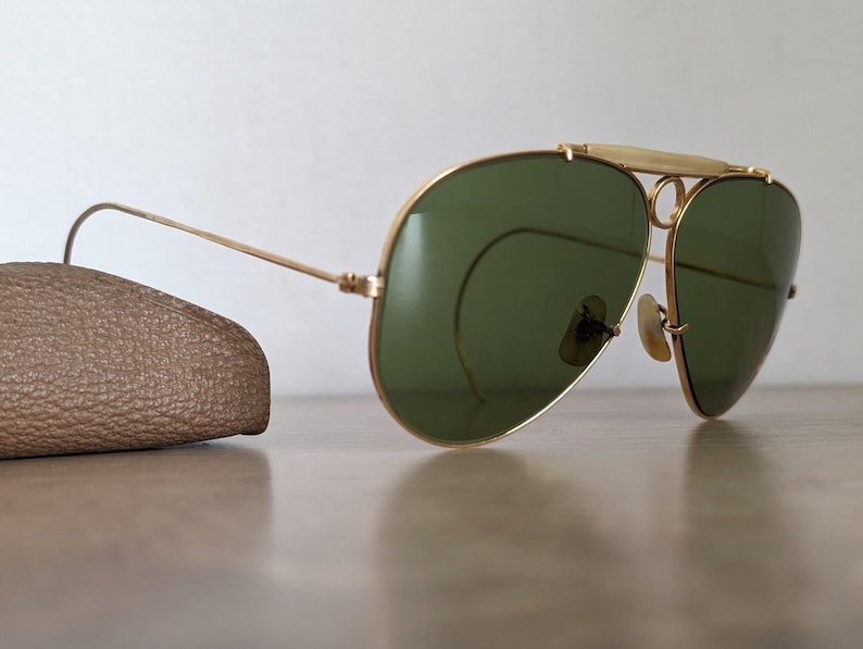 K&ouml;nnte beinhalten: Goldfarbene Aviator-Sonnenbrille mit gr&uuml;nen Gl&auml;sern. Die Sonnenbrille hat einen Doppelsteg und einen Stirnb&uuml;gel. Auf der linken Seite ist ein braunes Lederetui zu sehen. Die Sonnenbrille liegt auf einer hellbraunen Oberfl&auml;che.