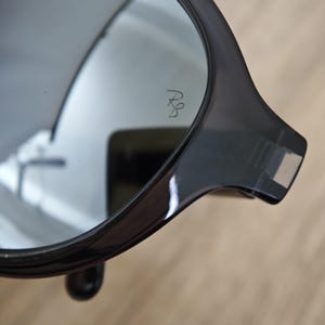 Puede incluir: Primer plano de gafas de sol gris oscuro con lente reflectante. Las gafas de sol tienen una montura redondeada y un peque&ntilde;o logotipo grabado en la lente. La montura es gruesa y brillante, con un peque&ntilde;o detalle redondeado cerca de la bisagra.