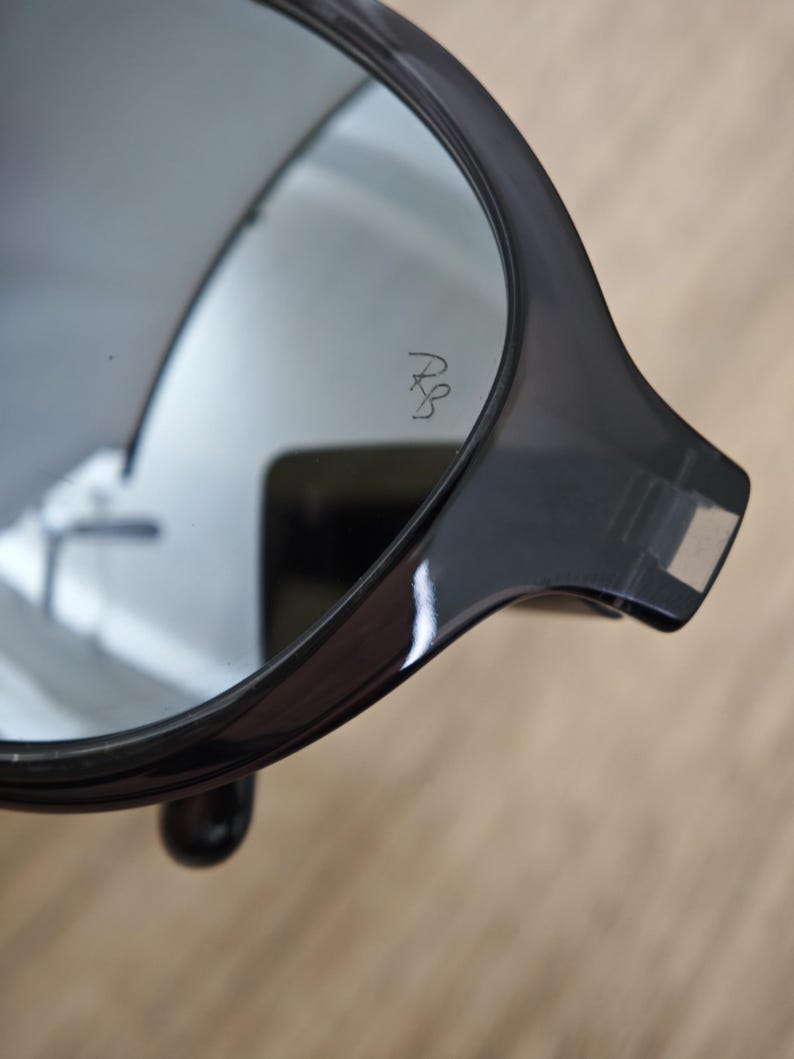 Puede incluir: Primer plano de gafas de sol gris oscuro con lente reflectante. Las gafas de sol tienen una montura redondeada y un peque&ntilde;o logotipo grabado en la lente. La montura es gruesa y brillante, con un peque&ntilde;o detalle redondeado cerca de la bisagra.