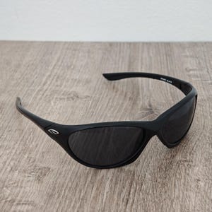 May include: Black sunglasses with a curved frame and dark lenses. The sunglasses have a sleek, sporty design. The text "Spark Black" is printed on the right temple. The sunglasses are on a wooden surface.