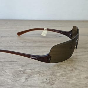 May include: Brown Versus sunglasses with a sleek, modern design. The sunglasses have a dark brown frame and lenses, with the brand name visible on the temple. The arms are a lighter brown color.
