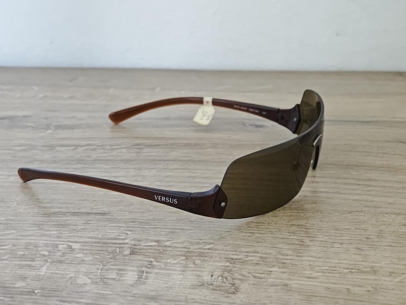 May include: Brown Versus sunglasses with a sleek, modern design. The sunglasses have a dark brown frame and lenses, with the brand name visible on the temple. The arms are a lighter brown color.
