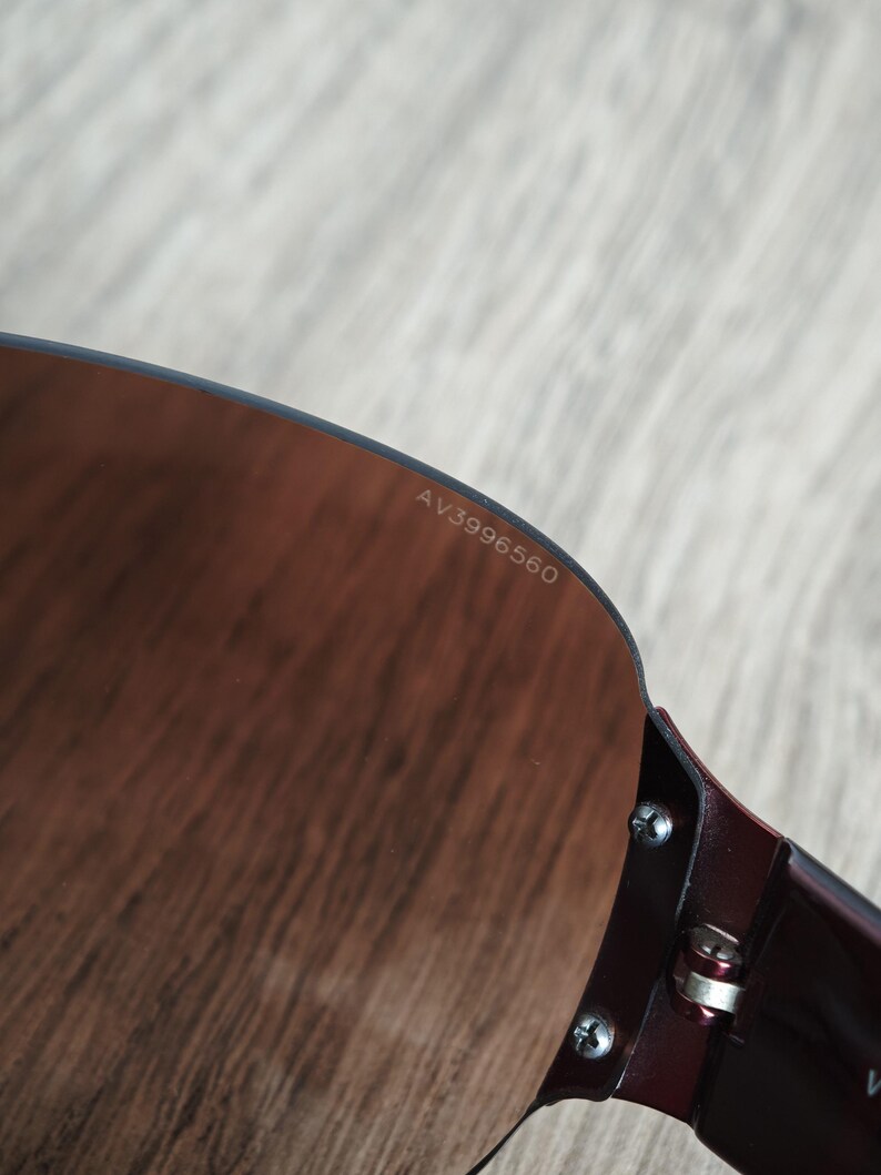 May include: Close-up of sunglasses with a dark burgundy frame and brown-tinted lenses. The text "AV3996560" is printed on the lens. The sunglasses are partially visible, showing the frame's hinge and screws.
