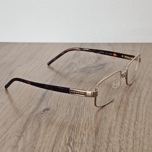 May include: Rectangular eyeglasses with a gold-colored frame and dark brown tortoise shell arms. The glasses are resting on a wooden surface. The brand name is visible on the arm.
