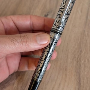 以下が含まれることがあります： 銀と黒の万年筆。装飾的な花柄のデザインが施されています。ペンにはシルバーのクリップと黒のキャップとペン先が付いています。木製の背景に、手でペンが持たれています。