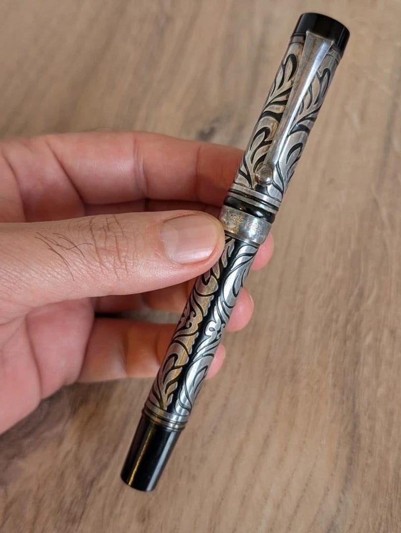 以下が含まれることがあります： 銀と黒の万年筆。装飾的な花柄のデザインが施されています。ペンにはシルバーのクリップと黒のキャップとペン先が付いています。木製の背景に、手でペンが持たれています。