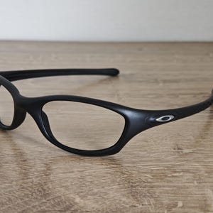 May include: Black eyeglasses with a sleek, modern design. The frames are matte black with a subtle silver logo on the temple. The lenses are clear, and the arms curve for a comfortable fit. The glasses are resting on a wooden surface.