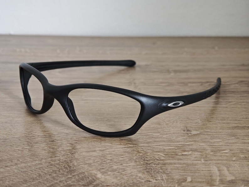 May include: Black eyeglasses with a sleek, modern design. The frames are matte black with a subtle silver logo on the temple. The lenses are clear, and the arms curve for a comfortable fit. The glasses are resting on a wooden surface.