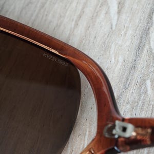Puede incluir: Primer plano de gafas de sol marrones con una lente oscura. La montura es de color marr&oacute;n transl&uacute;cido. El texto "KV7313993" est&aacute; impreso en el interior de la montura.