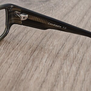 May include: Close-up of a pair of eyeglasses with dark frames. The text "Candidate 2.0" is printed on the temple. The glasses have a silver hinge and are set against a light brown wooden surface.