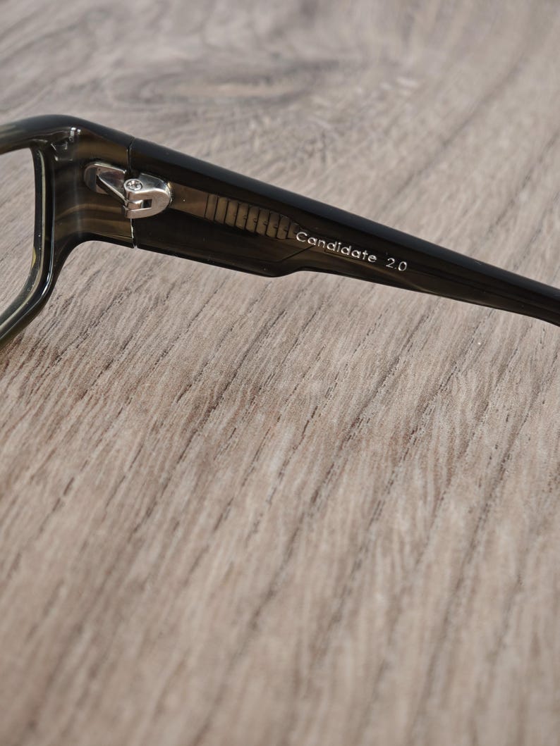 May include: Close-up of a pair of eyeglasses with dark frames. The text "Candidate 2.0" is printed on the temple. The glasses have a silver hinge and are set against a light brown wooden surface.