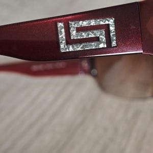 May include: Close-up of a pair of burgundy sunglasses. The frame features a silver geometric design on the temple. The lenses are a gradient brown color. The sunglasses are set against a neutral background.