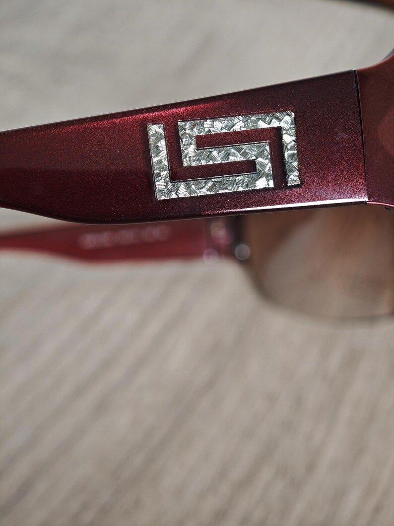 May include: Close-up of a pair of burgundy sunglasses. The frame features a silver geometric design on the temple. The lenses are a gradient brown color. The sunglasses are set against a neutral background.