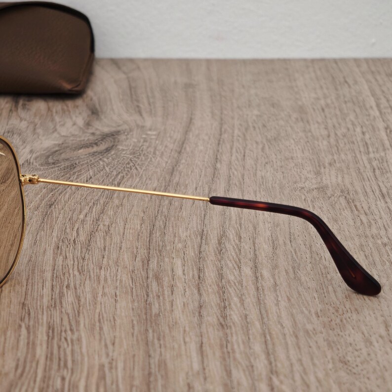 K&ouml;nnte beinhalten: Goldfarbene Aviator-Sonnenbrille mit braunen Gl&auml;sern. Die B&uuml;gel sind goldfarben mit dunkelbraunen Spitzen. Im Hintergrund befindet sich ein braunes Lederetui. Die Sonnenbrille liegt auf einer Holzoberfl&auml;che.