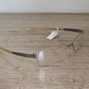 以下が含まれることがあります： 長方形のフレームと透明なレンズの眼鏡。 フレームは濃いグレーで、細くモダンなデザインです。 テンプルは薄いタン色です。 白いタグがメガネに付いています。