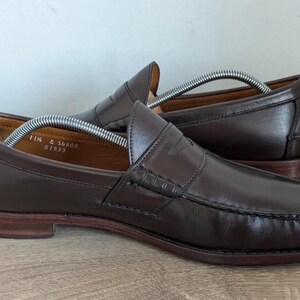 May include: A pair of brown leather penny loafers with a classic design. The shoes have a rounded toe, a penny strap across the vamp, and a stitched sole. The size is 11 1/2. The shoes are on a wooden surface.