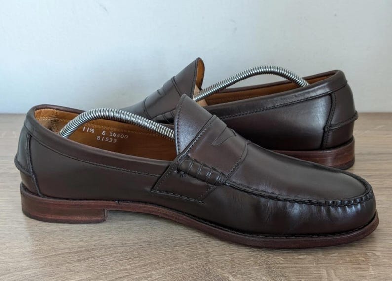 May include: A pair of brown leather penny loafers with a classic design. The shoes have a rounded toe, a penny strap across the vamp, and a stitched sole. The size is 11 1/2. The shoes are on a wooden surface.