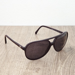 Puede incluir: Gafas de sol aviador de color púrpura oscuro con doble puente y lentes oscuras. Las gafas de sol tienen un pequeño logotipo plateado en la patilla. Sobre una superficie de madera clara.