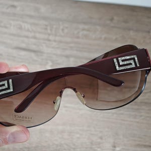May include: Dark burgundy sunglasses with gradient lenses. The frames feature a silver geometric design on the temples and the brand name "VERSACE" is visible on the lens. The sunglasses are held by a hand.
