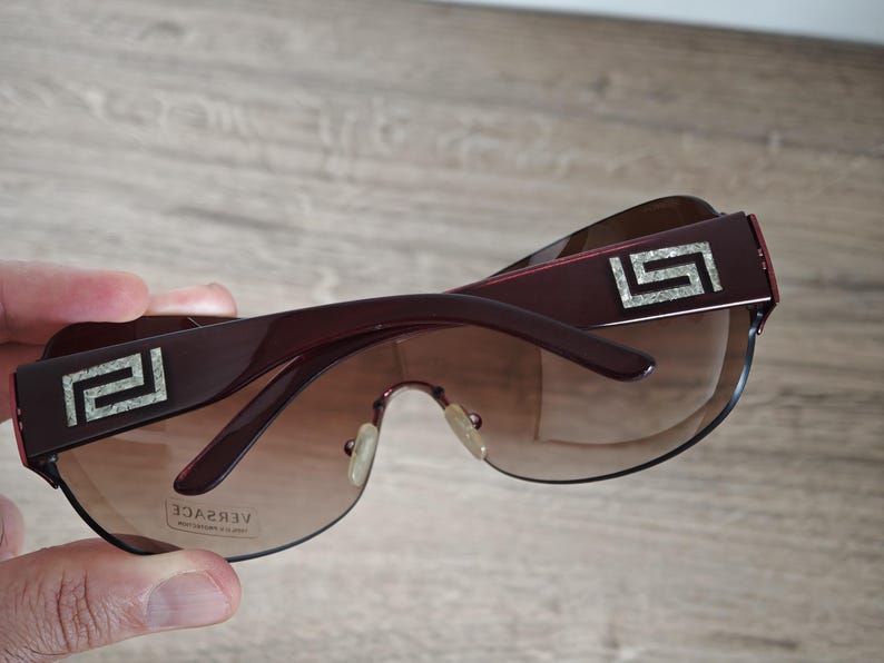 May include: Dark burgundy sunglasses with gradient lenses. The frames feature a silver geometric design on the temples and the brand name "VERSACE" is visible on the lens. The sunglasses are held by a hand.