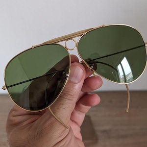 K&ouml;nnte beinhalten: Goldfarbene Aviator-Sonnenbrille mit gr&uuml;nen Gl&auml;sern. Die Sonnenbrille hat einen Doppelsteg und eine klassische Tropfenform. Die Rahmen sind goldfarben gl&auml;nzend, und die Gl&auml;ser sind dunkelgr&uuml;n. Die Sonnenbrille wird von einer Hand gehalten.