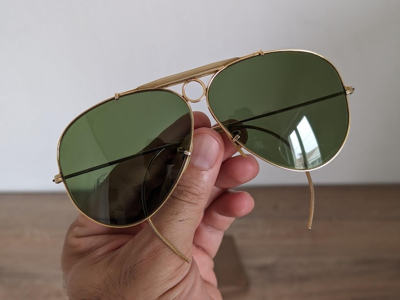 K&ouml;nnte beinhalten: Goldfarbene Aviator-Sonnenbrille mit gr&uuml;nen Gl&auml;sern. Die Sonnenbrille hat einen Doppelsteg und eine klassische Tropfenform. Die Rahmen sind goldfarben gl&auml;nzend, und die Gl&auml;ser sind dunkelgr&uuml;n. Die Sonnenbrille wird von einer Hand gehalten.
