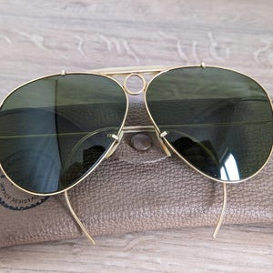 K&ouml;nnte beinhalten: Goldfarbene Aviator-Sonnenbrille mit gr&uuml;nen Gl&auml;sern auf einem braunen Lederetui. Die Sonnenbrille hat einen Doppelsteg und eine klassische Tropfenform. Das Etui hat einen Druckknopfverschluss und ein rundes Logo.