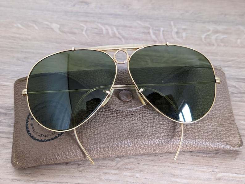 K&ouml;nnte beinhalten: Goldfarbene Aviator-Sonnenbrille mit gr&uuml;nen Gl&auml;sern auf einem braunen Lederetui. Die Sonnenbrille hat einen Doppelsteg und eine klassische Tropfenform. Das Etui hat einen Druckknopfverschluss und ein rundes Logo.