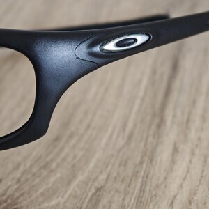 May include: Close-up of black eyeglasses with a white and silver logo on the temple. The glasses have a sleek, modern design and are set against a wooden background.