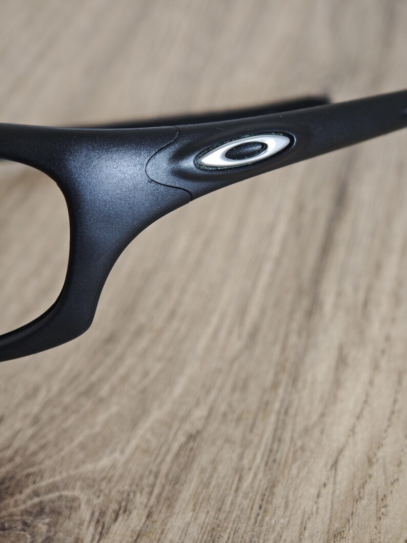 May include: Close-up of black eyeglasses with a white and silver logo on the temple. The glasses have a sleek, modern design and are set against a wooden background.