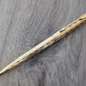 Könnte beinhalten: Ein goldfarbener Stift mit einem strukturierten, gravierten Muster. Der Stift hat ein schlankes, längliches Design mit einer Spitze. Der Clip und die Akzente des Stifts sind ebenfalls goldfarben. Der Stift liegt auf einer Holzoberfläche.