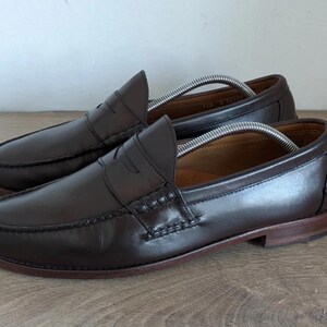 May include: A pair of dark brown leather penny loafers. The shoes have a classic design with a rounded toe, a strap across the vamp, and a low heel. Shoe trees are inserted inside the shoes.