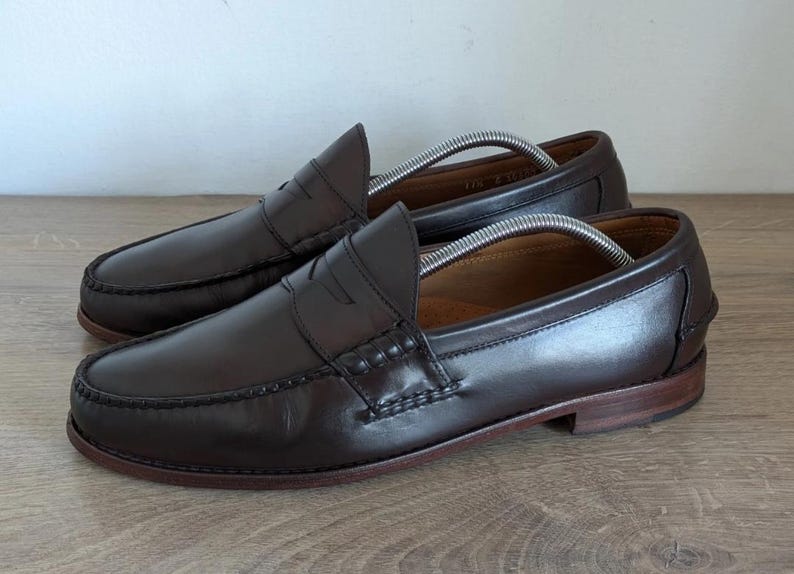 May include: A pair of dark brown leather penny loafers. The shoes have a classic design with a rounded toe, a strap across the vamp, and a low heel. Shoe trees are inserted inside the shoes.