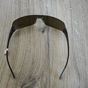 May include: Dark brown sunglasses with a curved, rimless design. The lenses are a dark, smoky color, and the arms are a darker shade of brown. The sunglasses are set against a wooden background.