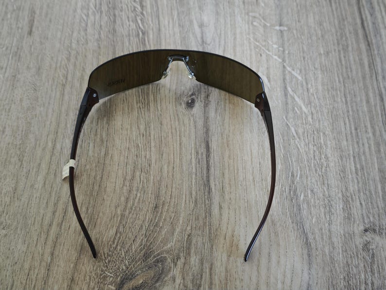 May include: Dark brown sunglasses with a curved, rimless design. The lenses are a dark, smoky color, and the arms are a darker shade of brown. The sunglasses are set against a wooden background.