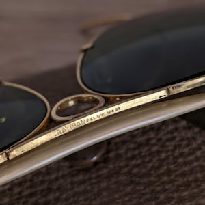 K&ouml;nnte beinhalten: Nahaufnahme einer Ray-Ban Sonnenbrille mit goldenem Gestell und dunklen Gl&auml;sern. Der Text "RAY-BAN B&L 1/10 12K GF" ist auf dem B&uuml;gel aufgedruckt. Die Sonnenbrille ist teilweise sichtbar und zeigt die Details von Gestell und Gl&auml;sern.