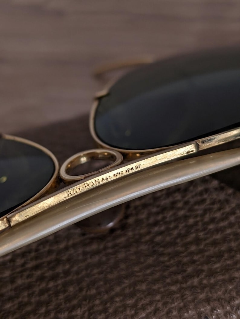 K&ouml;nnte beinhalten: Nahaufnahme einer Ray-Ban Sonnenbrille mit goldenem Gestell und dunklen Gl&auml;sern. Der Text "RAY-BAN B&L 1/10 12K GF" ist auf dem B&uuml;gel aufgedruckt. Die Sonnenbrille ist teilweise sichtbar und zeigt die Details von Gestell und Gl&auml;sern.