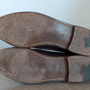 May include: Close-up of the soles of two brown leather shoes. The soles are worn, showing signs of use. The stitching is visible around the edges. The heels are a darker brown color.