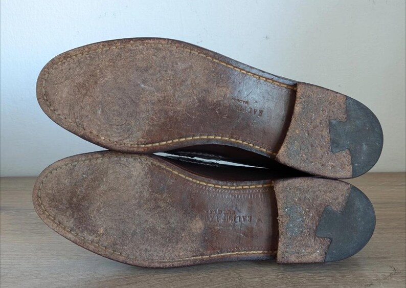 May include: Close-up of the soles of two brown leather shoes. The soles are worn, showing signs of use. The stitching is visible around the edges. The heels are a darker brown color.