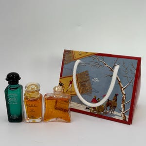 Può includere: Tre bottiglie di profumo di varie forme e colori, tra cui verde, ambra e trasparente. È presente anche un sacchetto regalo decorativo con una scena invernale e un manico in corda bianca.