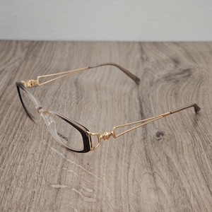 Op de afbeelding: Versace bril met een donkerbruin en transparant verloopframe en dunne goudkleurige metalen pootjes. De scharnieren zijn versierd met een klein gouden Medusa-hoofd embleem. Het woord Versace staat in het zwart op de rechter lens.