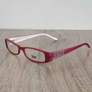 May include: A pair of rectangular, burgundy-colored eyeglasses with the brand name "D&G" printed on the left lens. The right temple arm features the brand name in rhinestones. The arms are two-toned, with a light pink interior and white tips.