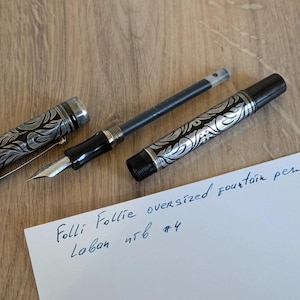 以下が含まれることがあります： シルバーと黒のデザインの特大万年筆。キャップとバレルには装飾的な模様があります。ペンは分解されており、ペン先とインクカートリッジが見えます。紙には「Folli Follie oversized fountain pen」と書かれています。