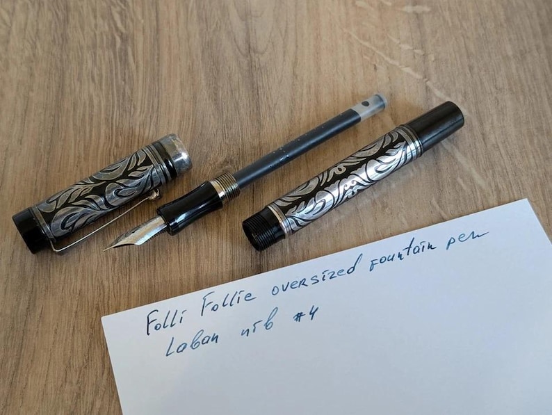 以下が含まれることがあります： シルバーと黒のデザインの特大万年筆。キャップとバレルには装飾的な模様があります。ペンは分解されており、ペン先とインクカートリッジが見えます。紙には「Folli Follie oversized fountain pen」と書かれています。