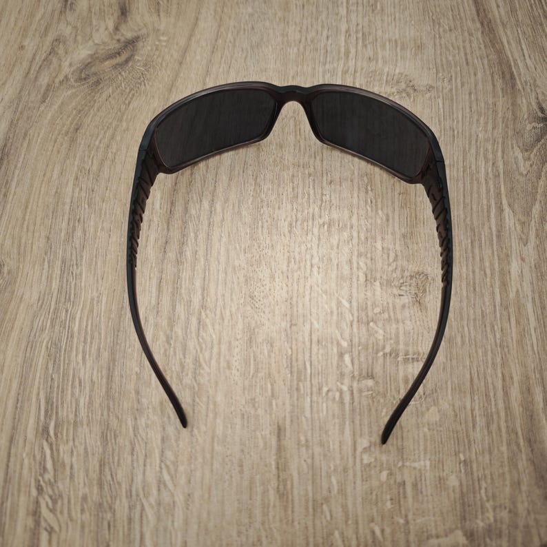 Puede incluir: Un par de gafas de sol de color marr&oacute;n oscuro con lentes rectangulares y patillas texturizadas. La montura presenta un acabado mate s&oacute;lido con un puente curvo que conecta las dos lentes. Las patillas son ligeramente c&oacute;nicas.