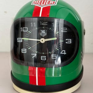 以下が含まれることがあります： 緑色のレーシングヘルメット型置き時計。上部と中央に赤い縦線とTAG Heuerのロゴが入っています。文字盤は黒で、1から12までの白い数字、白い針、そして前面を覆う透明なバイザーが付いています。