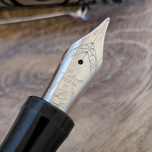 以下が含まれることがあります： 「Laban」のブランド名が刻まれた銀色の万年筆のペン先をクローズアップ。ペン先には装飾的な装飾と「IRIDIUM」の文字が施されています。ペンのボディは黒く光沢があります。