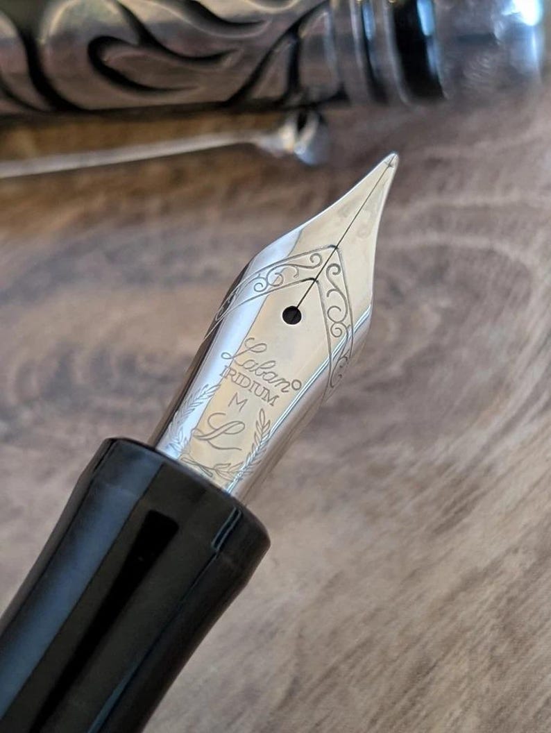 以下が含まれることがあります： 「Laban」のブランド名が刻まれた銀色の万年筆のペン先をクローズアップ。ペン先には装飾的な装飾と「IRIDIUM」の文字が施されています。ペンのボディは黒く光沢があります。